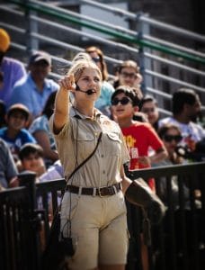 A woman emcee speaks to a crowd in bleachers, pointing and engaging during an outdoor event.